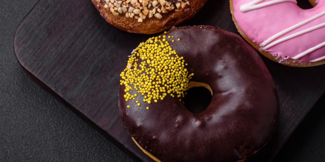 Three donuts on a dark wooden surface: one with yellow sprinkles, one pink with white drizzle, and one with chopped nuts.