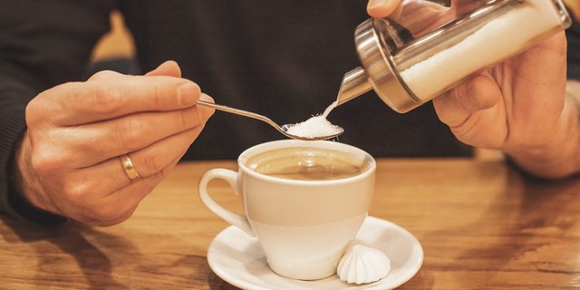 A person adding a spoonful of sugar to a cup of coffee on a wooden table